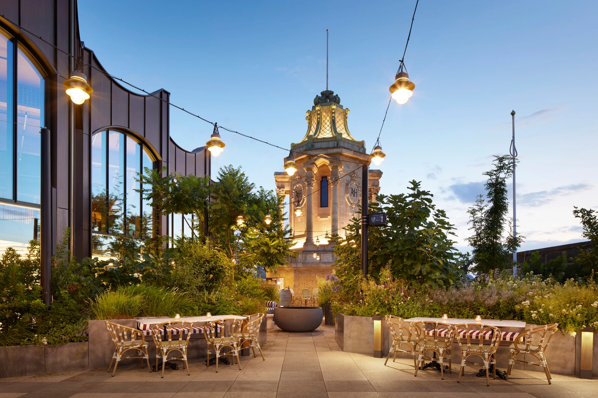 The Arding Room roof garden at dusk, festoon lights over café tables and the Arding and Hobbs clock tower